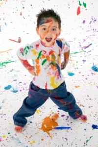 young boy covered in rainbow paint splotches in from of white background with rainbow paint splotches