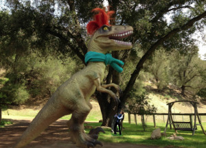 T-Rex statue wearing green scarf and red feather on head in park next to a person