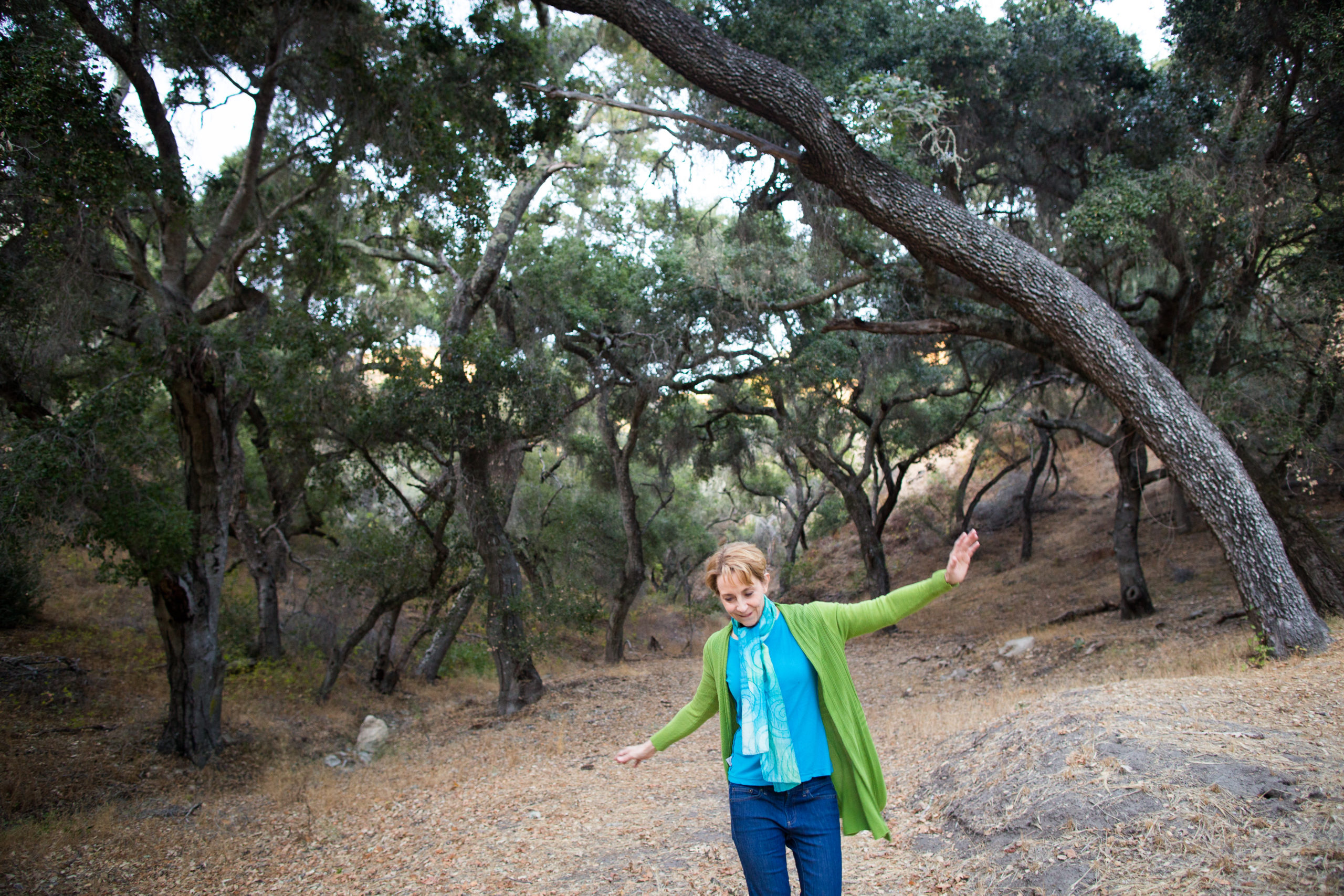 Martha Beck in bright blue top and green cardigan dancing in the woods