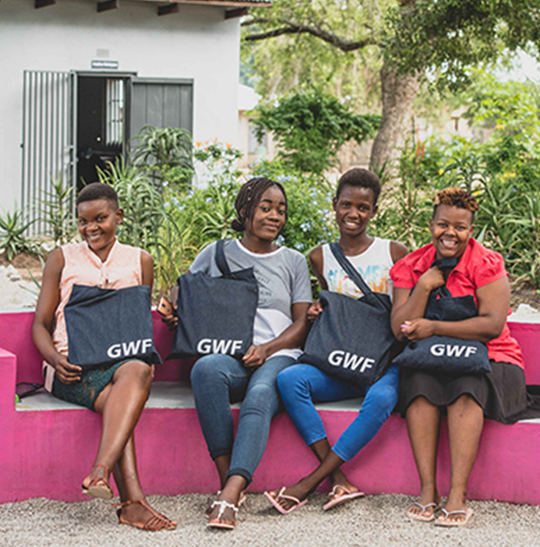 Four young smiling women with GWF tote bags sitting on purple bench outside