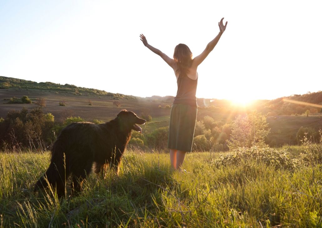 woman with arms flung wide standing in nature with dog at golden hour