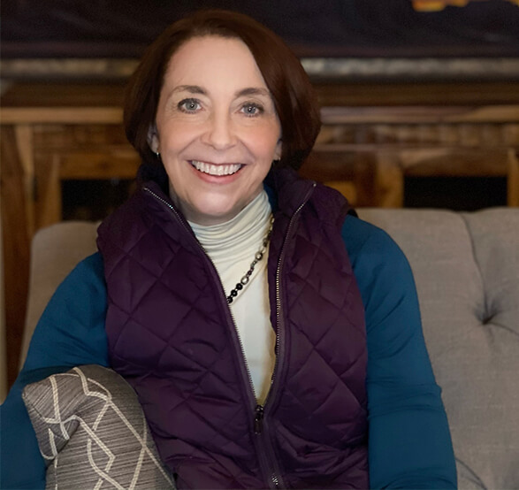 Martha Beck in a purple vest sitting on a gray couch smiling at the camera