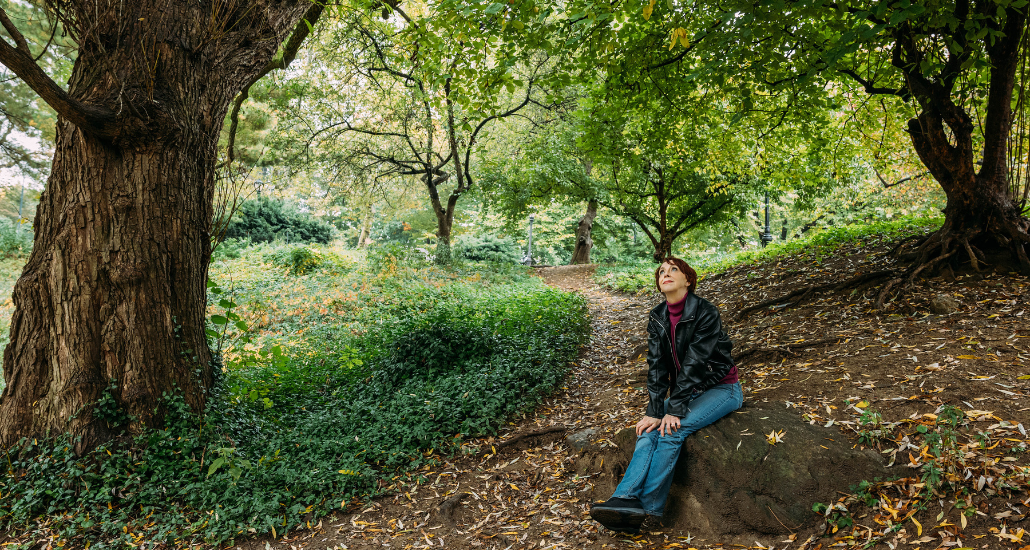Martha sitting on a large rock looking reflective underneath a canopy of green leaves.