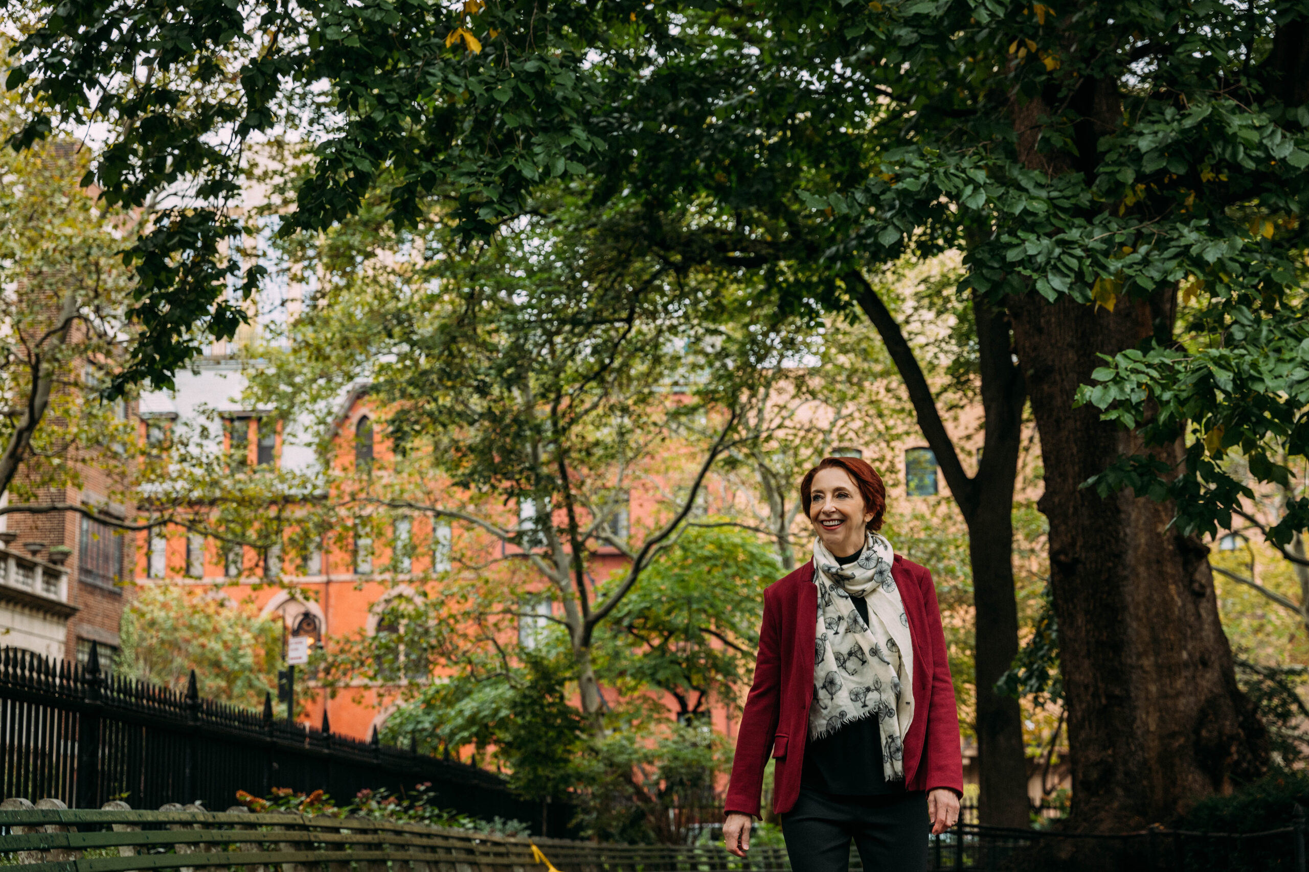 Martha walking in a red coat with a white and green scarf on, under a canopy of trees with buildings in the distance.