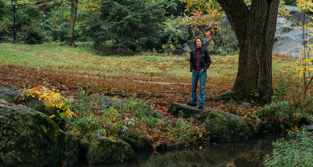 Martha in a leather jacket standing amidst fallen leaves under a tree beside a stream.